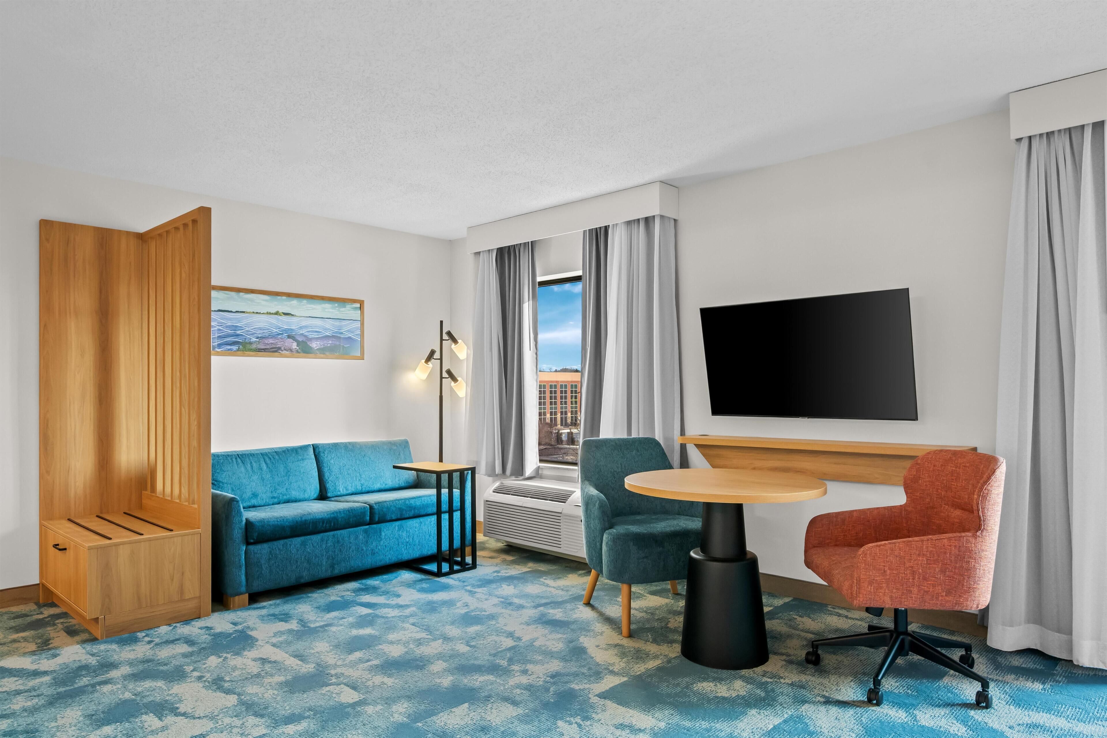 Habitación de hotel con sofá azul, aparador de madera, mesa redonda, dos sillas, televisor y ventana con cortinas.