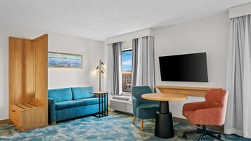 Habitación de hotel con sofá azul, aparador de madera, mesa redonda, dos sillas, televisor y ventana con cortinas.