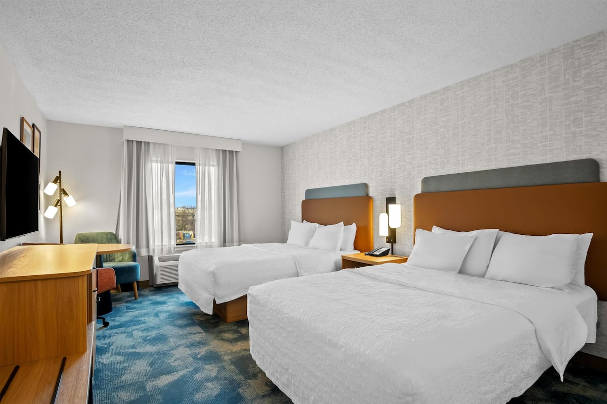 A hotel room with two beds, a desk, a chair, a TV, and a window with curtains.