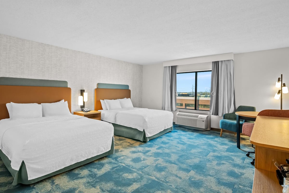 A hotel room with two beds, a desk, a chair, and a large window with curtains.