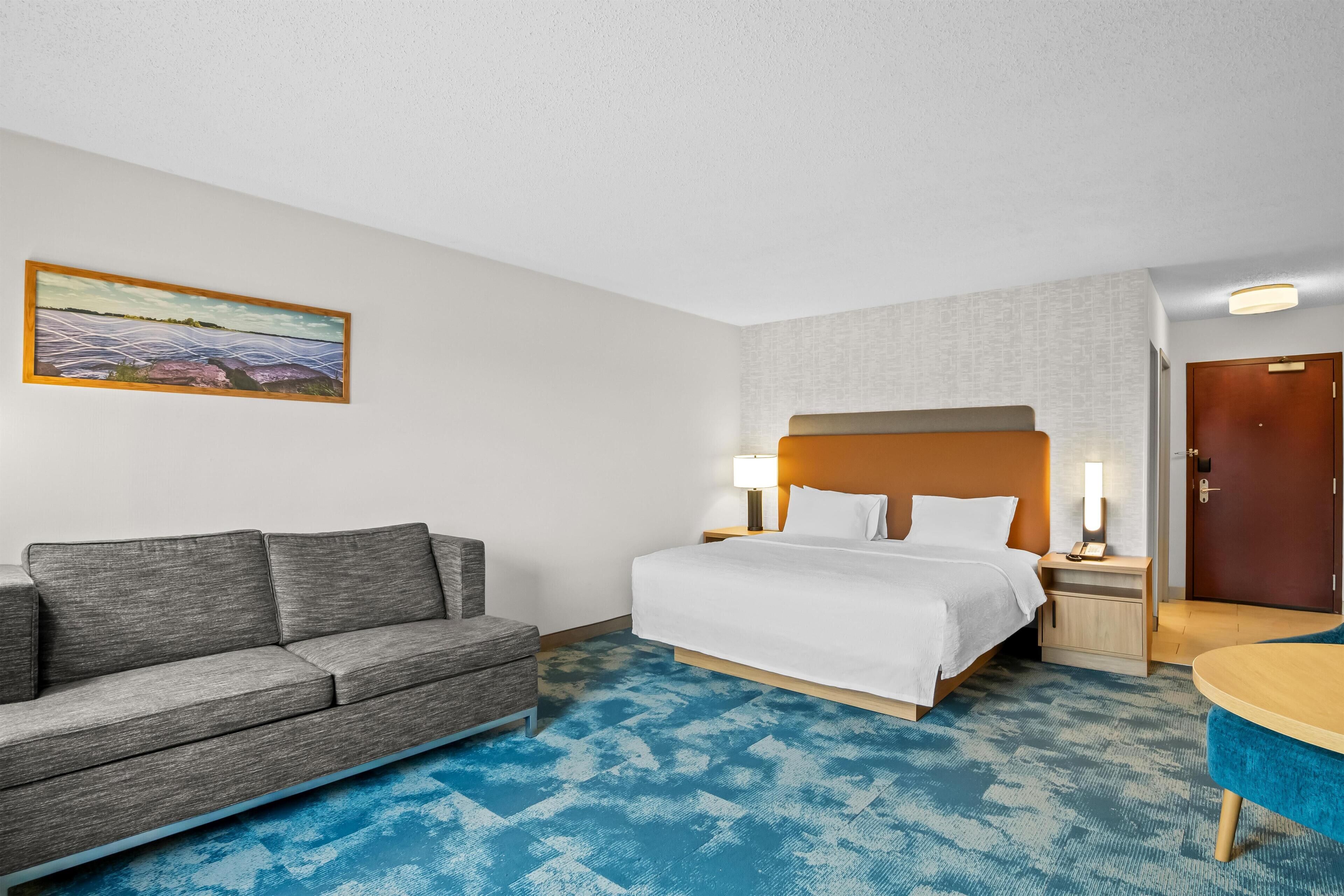 A hotel room with a bed, a sofa, a desk, and a painting on the wall.