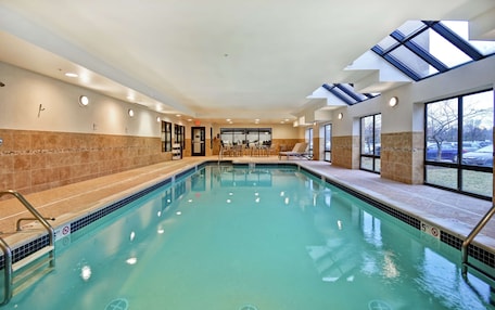 An indoor swimming pool with clear water, a sunroof, and a seating area with tables and chairs.