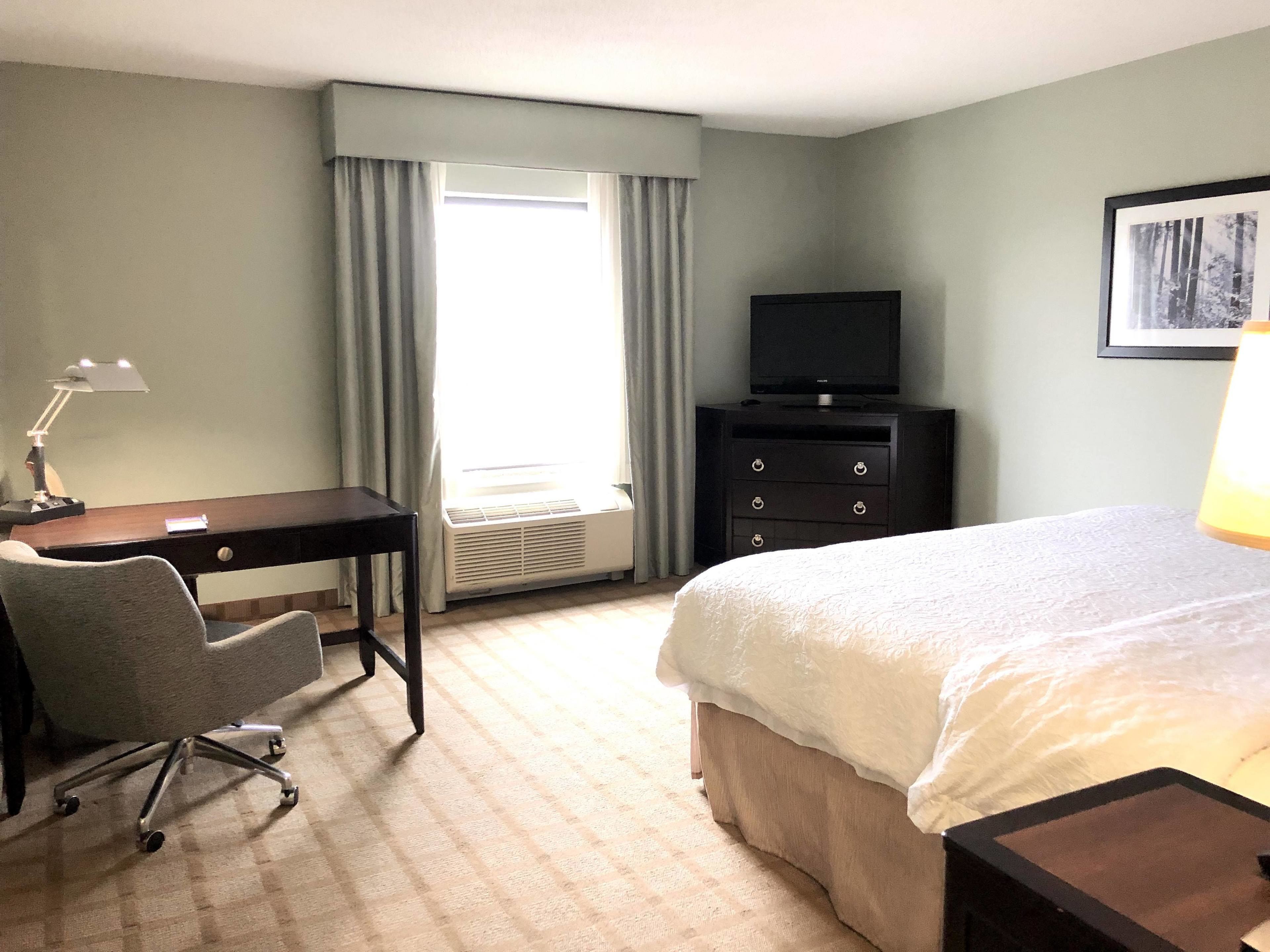 Una habitación de hotel con cama, escritorio con silla, televisor y ventana con cortinas.