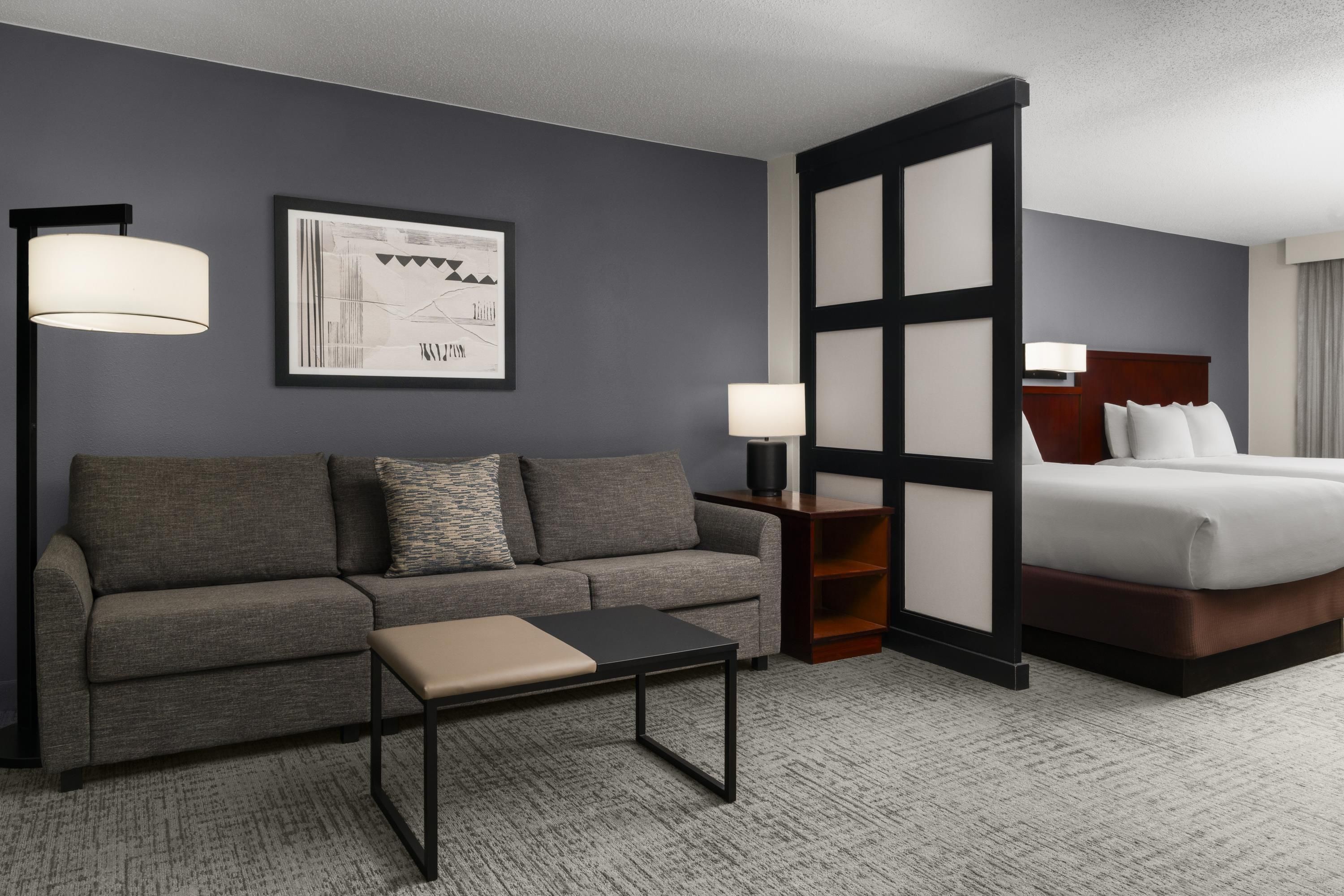 A hotel room with a grey sofa, a small table, a bed with white linens, a nightstand with a lamp, and a framed picture on the wall.