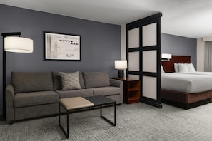 A hotel room with a grey sofa, a small table, a bed with white linens, a nightstand with a lamp, and a framed picture on the wall. - Hyatt Select Richmond Airport (Richmond)