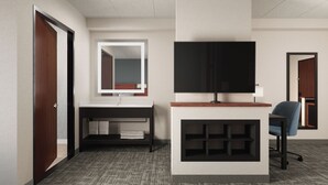 A hotel room with a wooden door, a bathroom with a mirror, a flat-screen TV, a desk, and a chair.