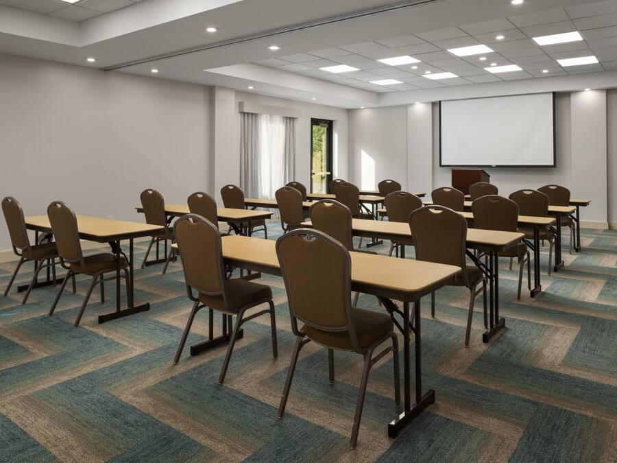 A conference room with multiple tables and chairs, carpeted floor, and a projector screen on the wall.