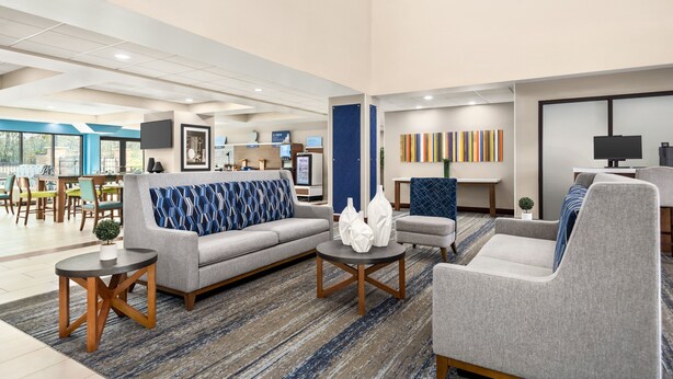 Un hall d’hôtel moderne avec un canapé gris, des fauteuils, une table basse et, au fond, un coin repas.