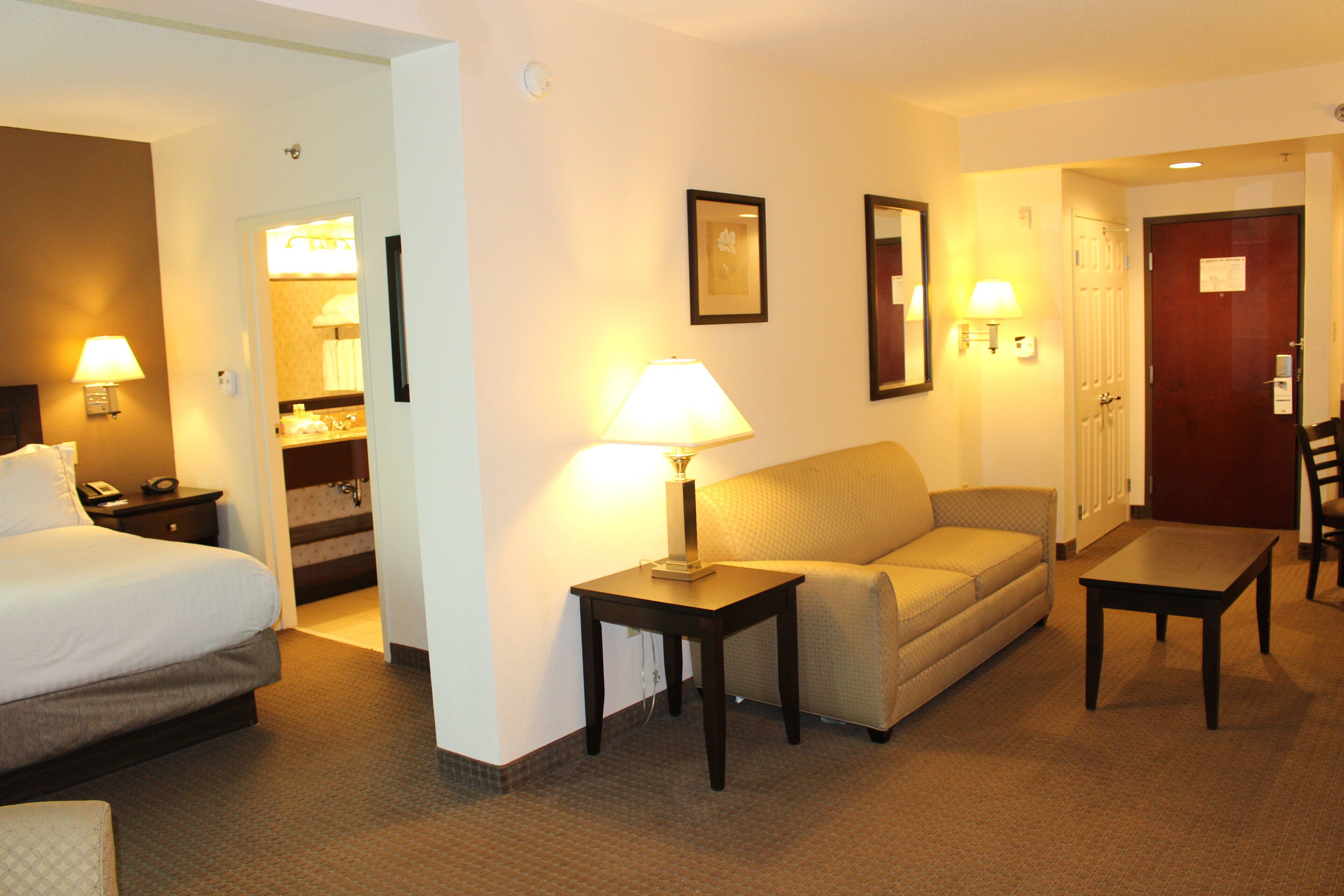 A hotel room with a bed, a sofa, a table, a lamp, a mirror, and a door.