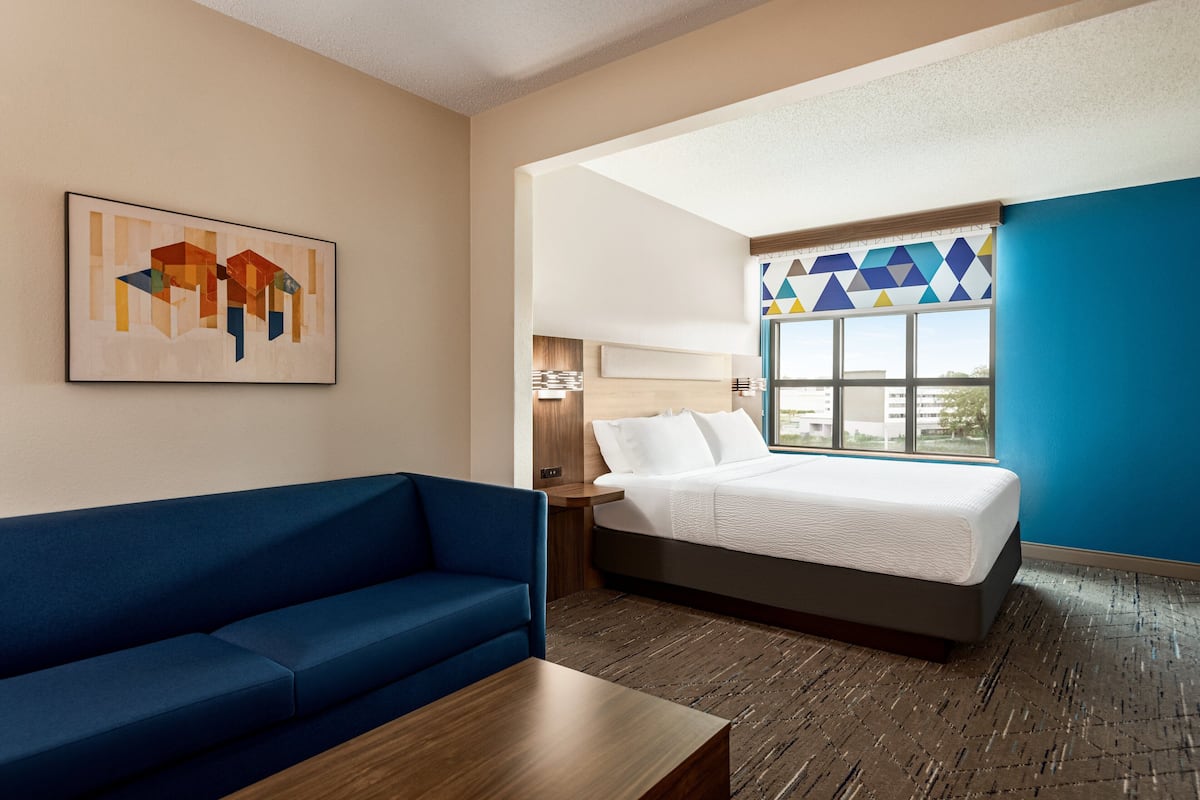 Una habitación de hotel con una cama, un sofá azul, una mesa de centro de madera, una lámpara de pared y una obra de arte geométrica en la pared.