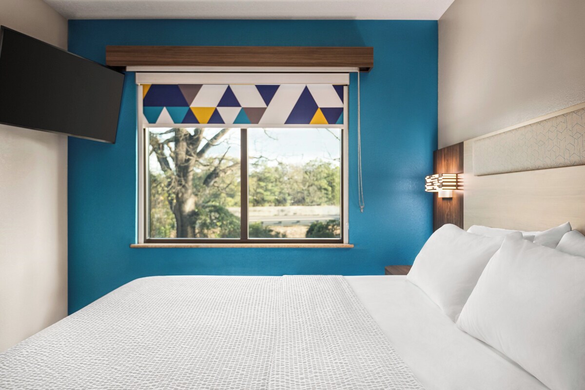 Une chambre d’hôtel avec un lit, une fenêtre ornée d’un rideau à motifs géométriques, et une vue sur des arbres et une route à l’extérieur.