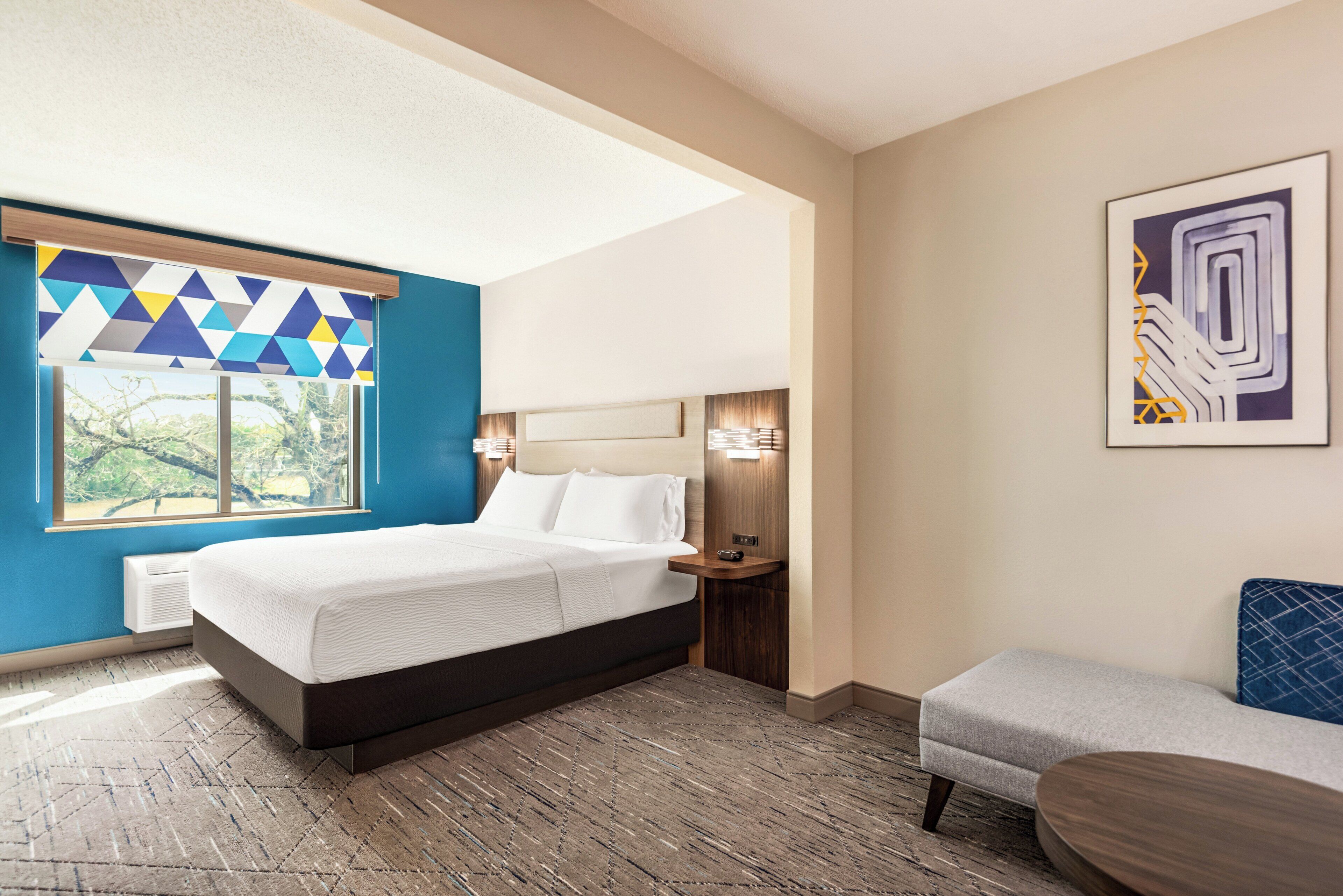 A modern hotel room with a large bed, a small seating area, and a window with geometric blinds.