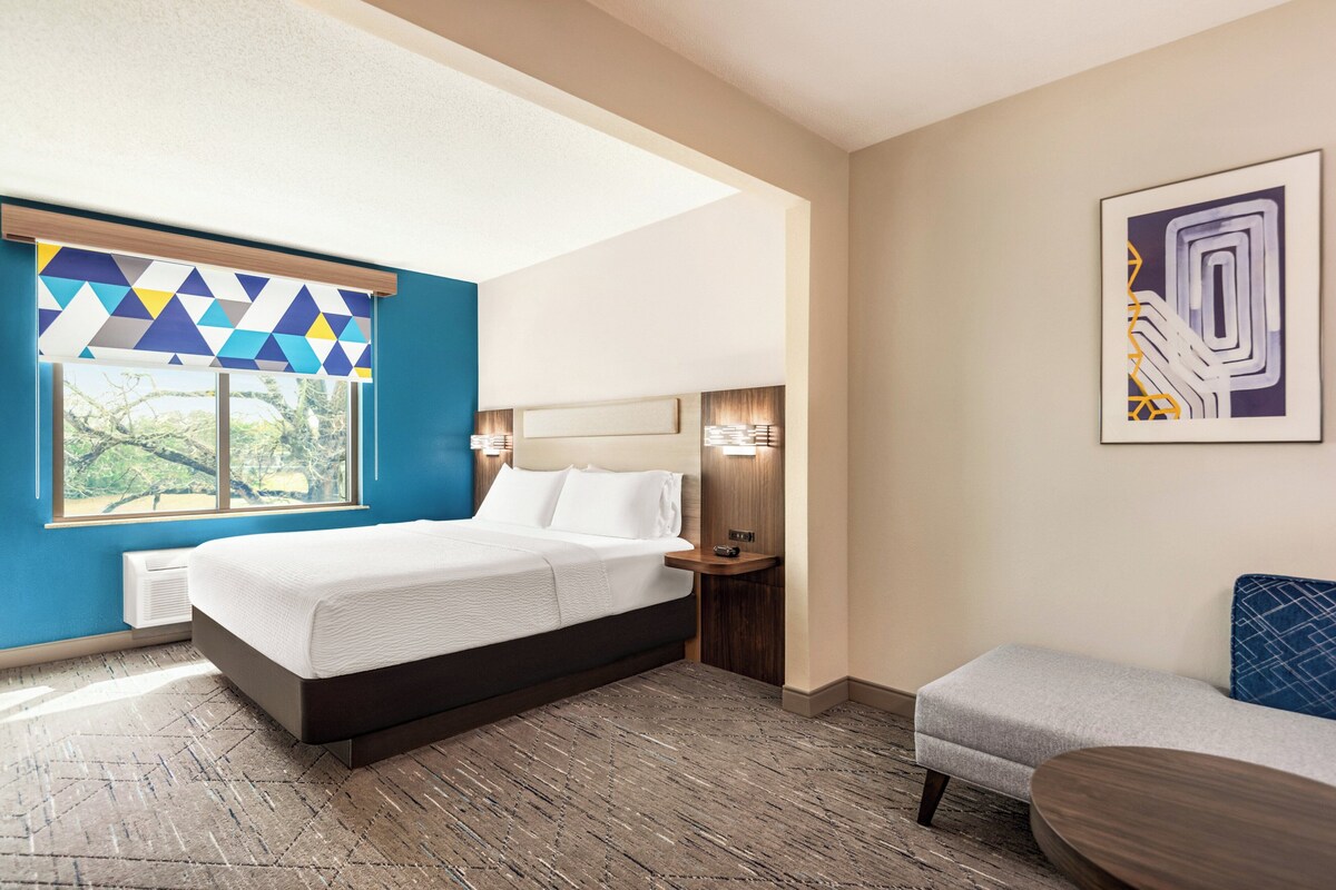 Une chambre d’hôtel moderne dotée d’un grand lit, d’un petit coin salon et d’une fenêtre avec des stores à motifs géométriques.