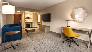 Une chambre d’hôtel avec un canapé bleu, un fauteuil de bureau jaune, un bureau en bois, un téléviseur à écran plat et une lampe moderne.