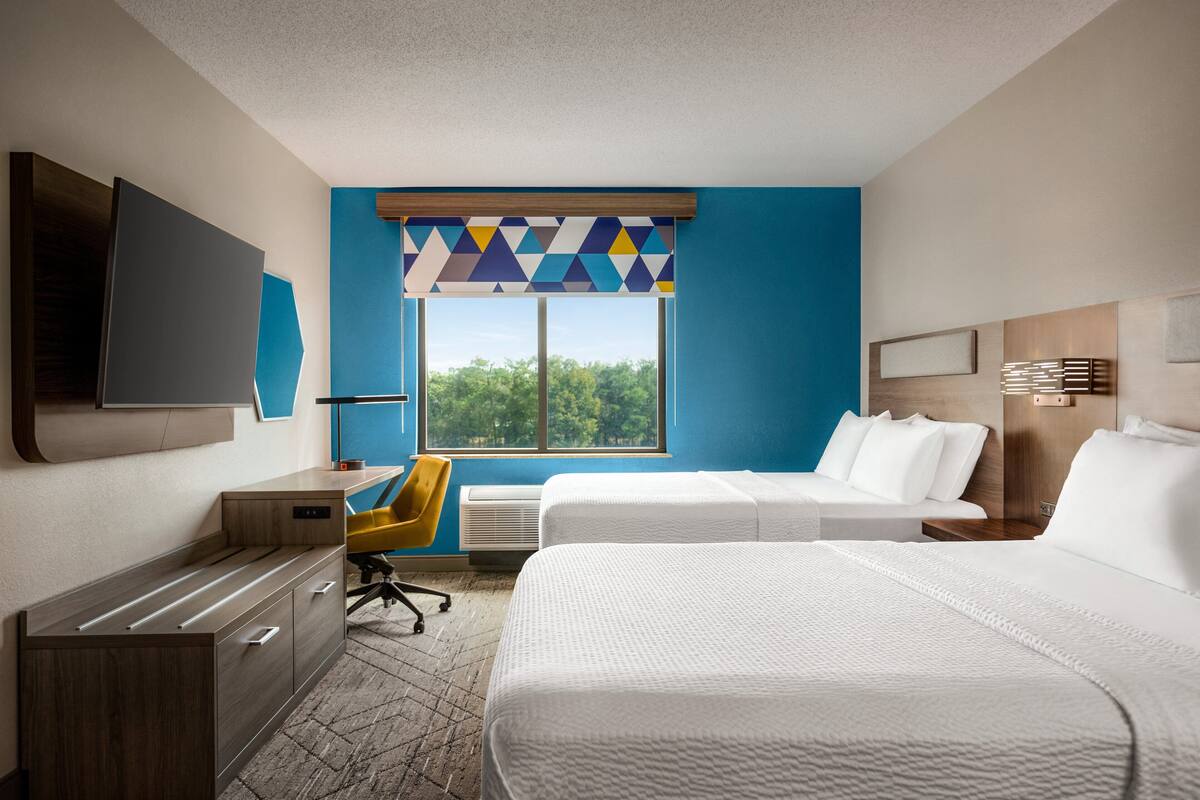 Habitación de hotel con escritorio, dos camas, televisor y ventana con vista a árboles.