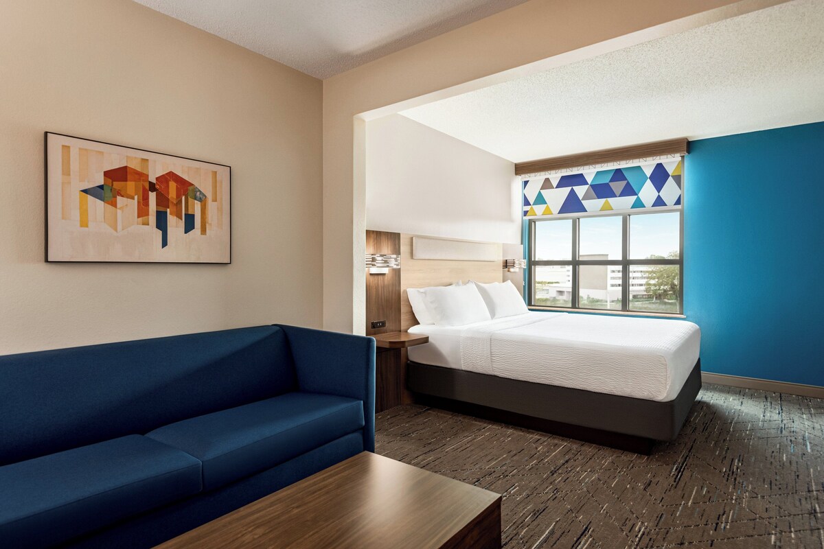 Une chambre d’hôtel avec un lit, un canapé bleu, une table basse en bois, une lampe fixée au mur et une œuvre d’art géométrique accrochée au mur.