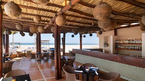 An outdoor seating area with wicker furniture, a bar with a tiled backsplash, and a view of the beach. - Grand Decameron Los Cabos, A Trademark  Resort (San José del Cabo)