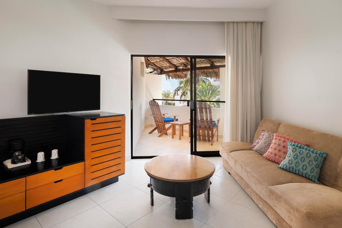 A modern living room with a flat-screen TV, a wooden coffee table, a sofa with colorful cushions, and a sliding glass door leading to an outdoor patio.