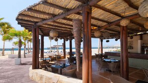 A beachside patio with wicker furniture, a stone wall, and a thatched roof structure. - Grand Decameron Los Cabos, A Trademark  Resort (San José del Cabo)