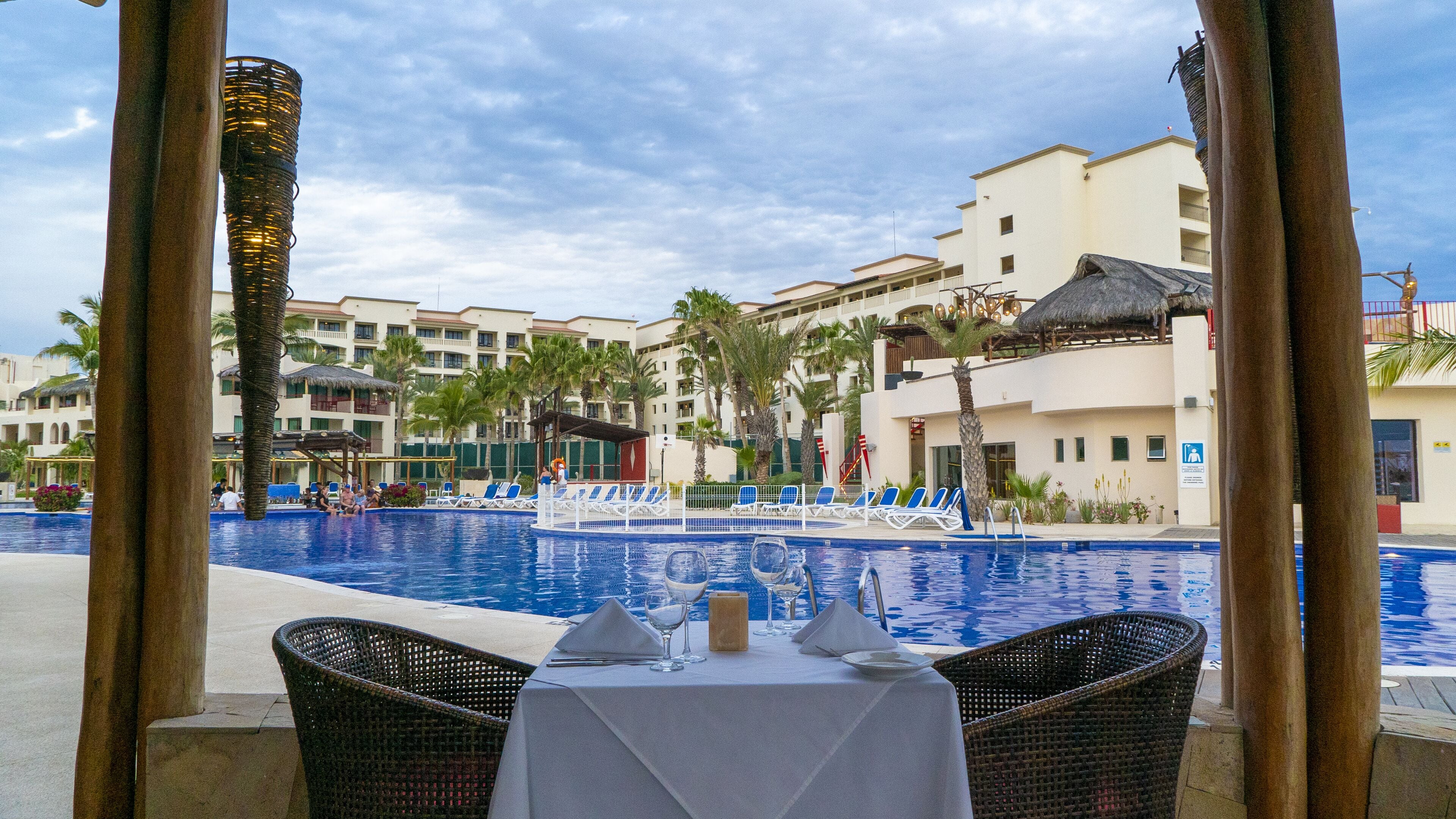 Un espace de restauration aménagé au bord de la piscine, avec une table dressée pour deux personnes, donnant sur une piscine, des chaises longues et des palmiers.