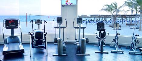Uma academia com aparelhos de cardio e uma tela de TV, com vista para uma praia com cadeiras de sol.