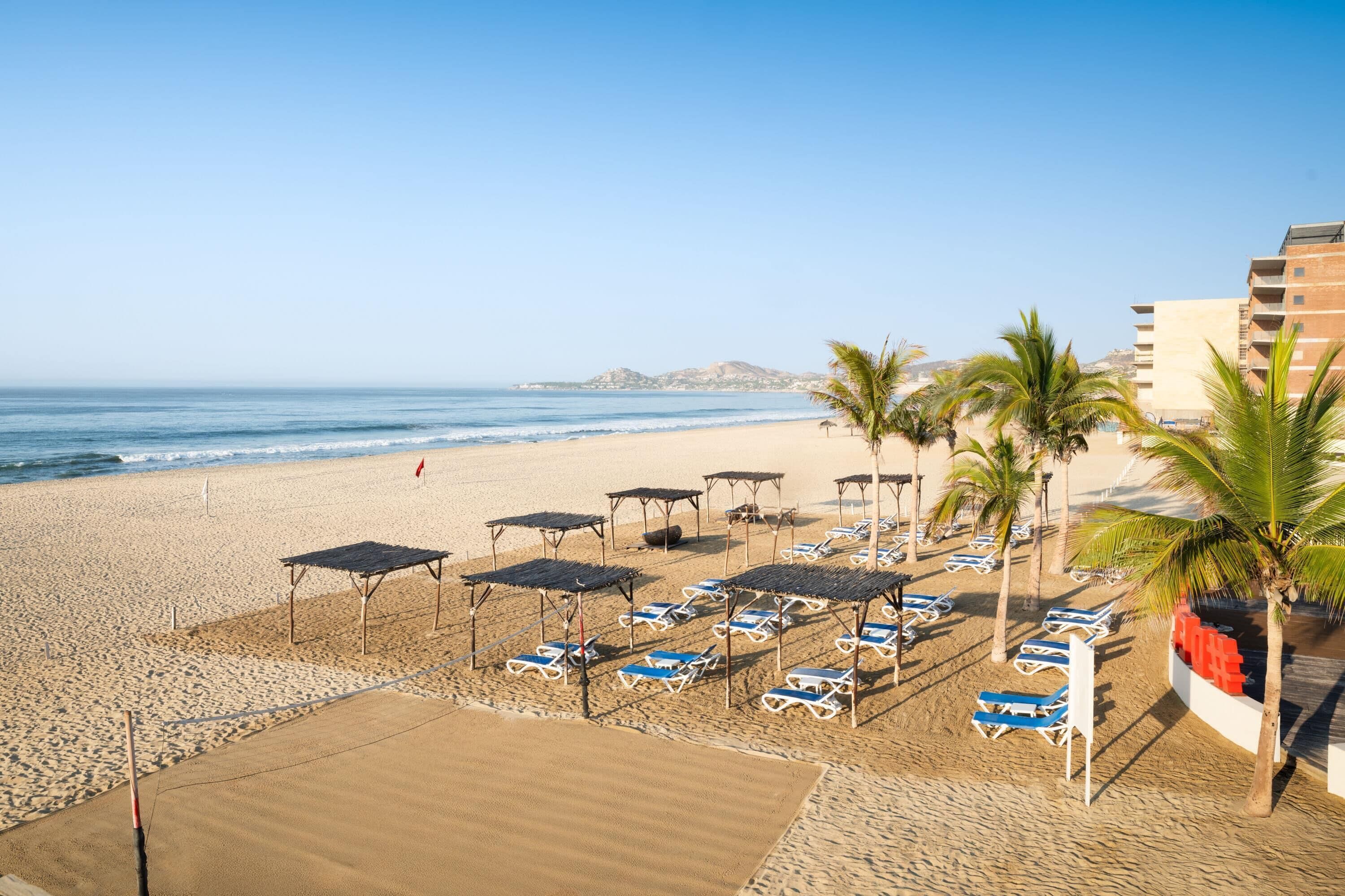 A beach with sun loungers, umbrellas, and palm trees.