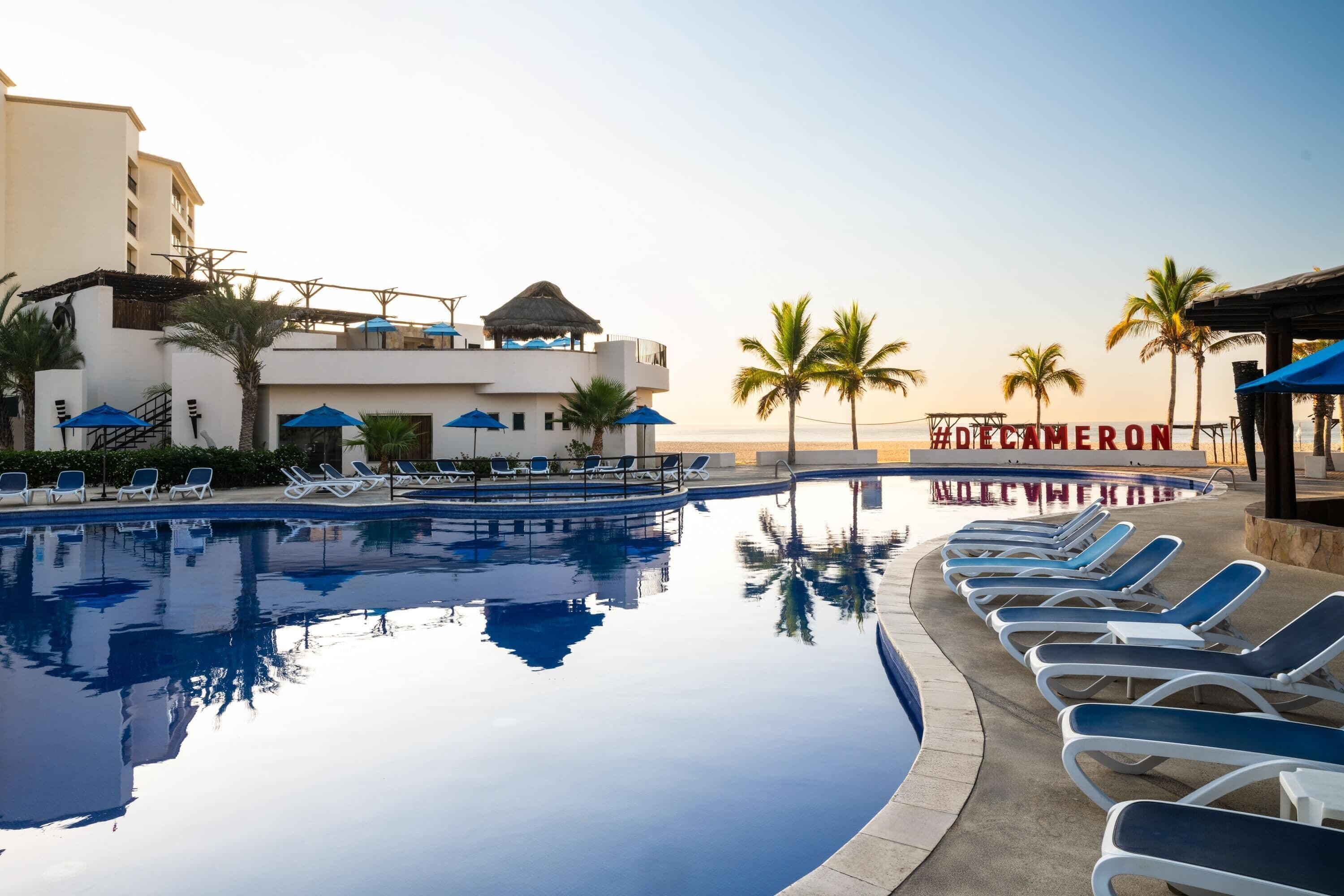 A swimming pool with lounge chairs and umbrellas, a building with a sign reading 'HOTEL MEROR,' and palm trees.