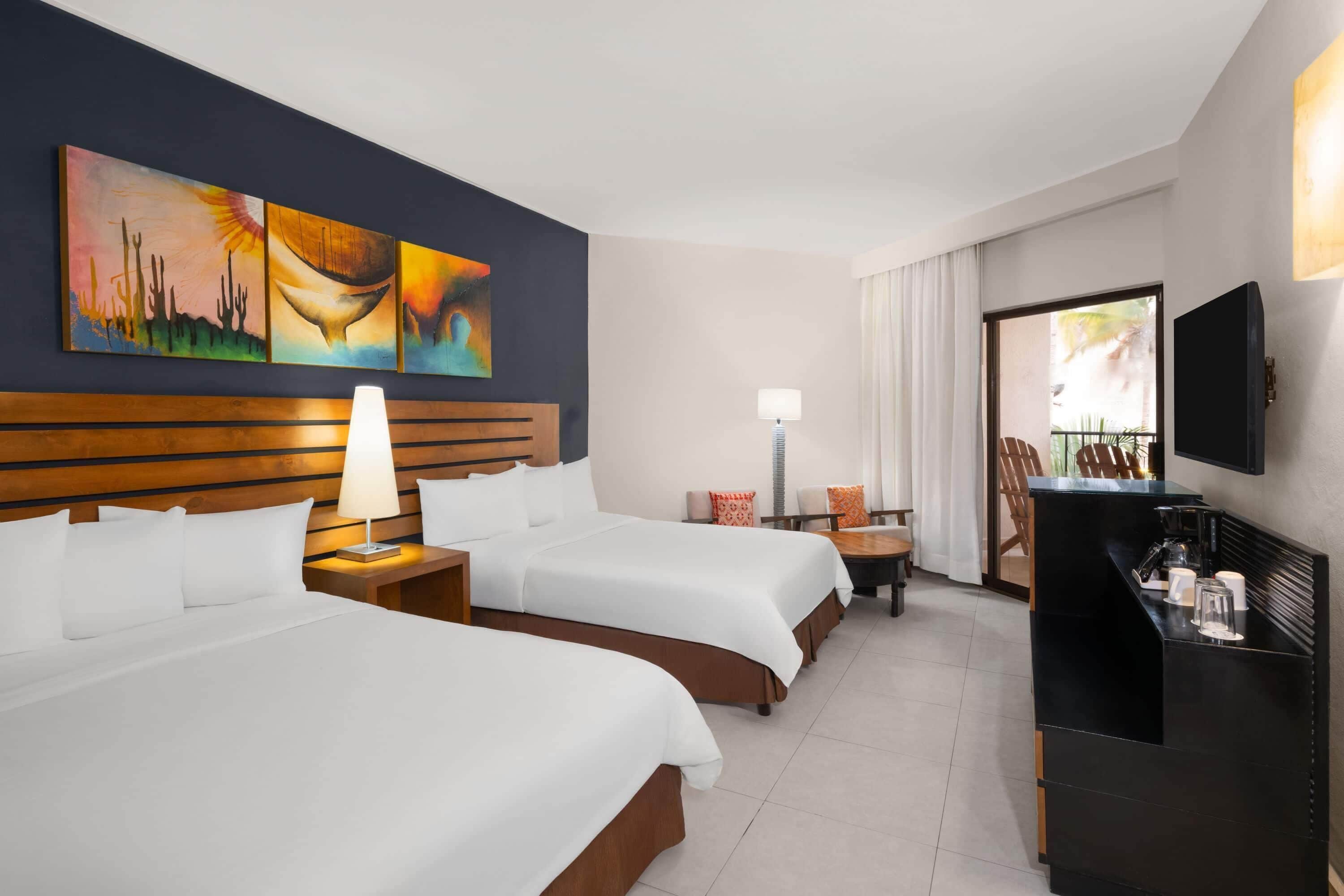 A hotel room with two beds, a desk, a chair, a TV, artwork on the wall, and a window.