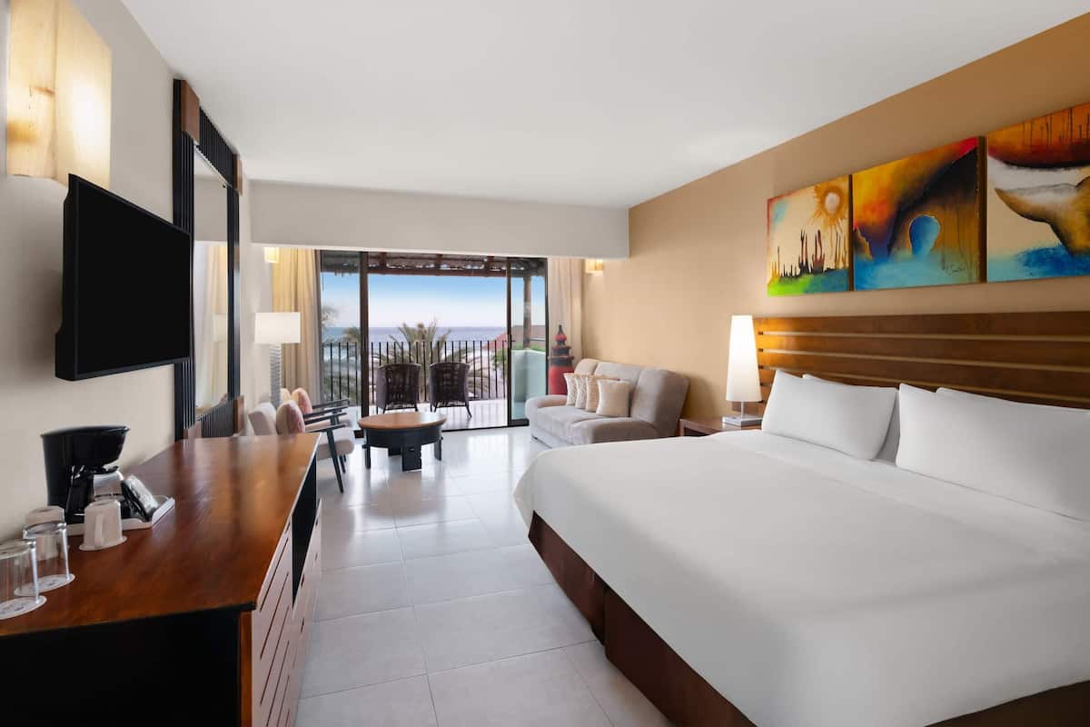 A hotel room with a large bed, a desk with a coffee maker, a TV, a balcony with a view, and artwork on the wall.