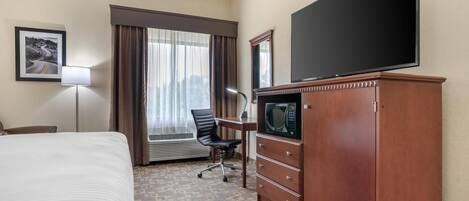 A hotel room with a bed, a desk with a flat-screen TV, a microwave, a chair, and a large window with curtains.