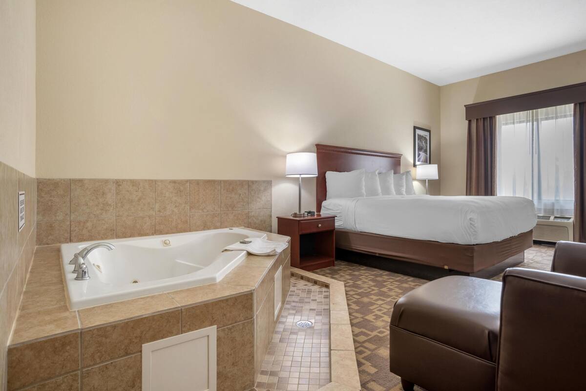 Habitación de hotel con una bañera grande, una cama con cabecera, una mesita de noche, una lámpara y una ventana con cortinas.