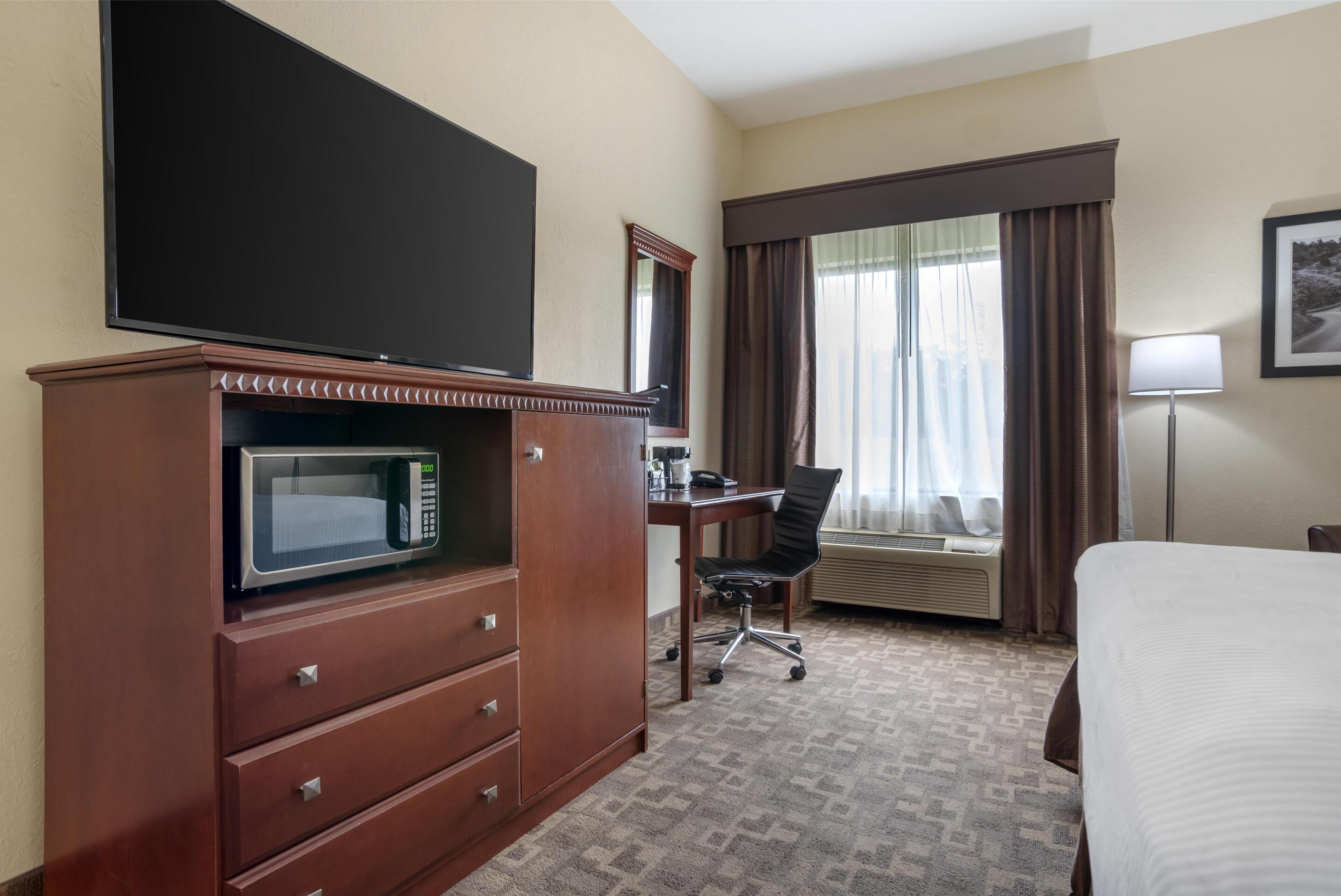 Une chambre d’hôtel comprenant un lit, un bureau avec une chaise, un téléviseur à écran plat, un four à micro-ondes, une commode, un appareil de climatisation et une fenêtre avec des rideaux.