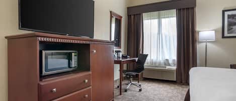 A hotel room with a bed, a desk with a chair, a flat-screen TV, a microwave, a dresser, an air conditioning unit, and a window with curtains.