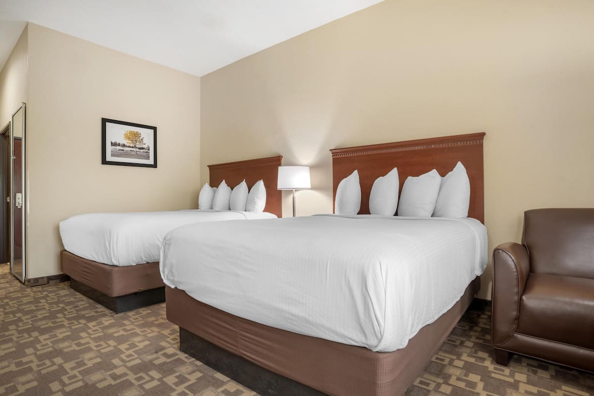 Habitación de hotel con dos camas, un sillón de cuero marrón, un cuadro enmarcado en la pared y una alfombra estampada.