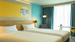 A hotel room with two beds, a wooden headboard, a small table, a chair, and a window with curtains. - B&B HOTEL Warrington (Warrington)
