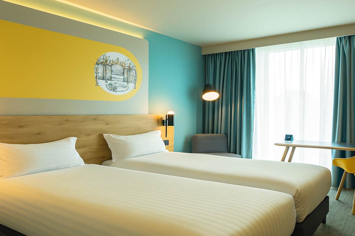 Een hotelkamer met twee bedden, een houten hoofdeinde, een klein tafeltje, een stoel en een raam met gordijnen.