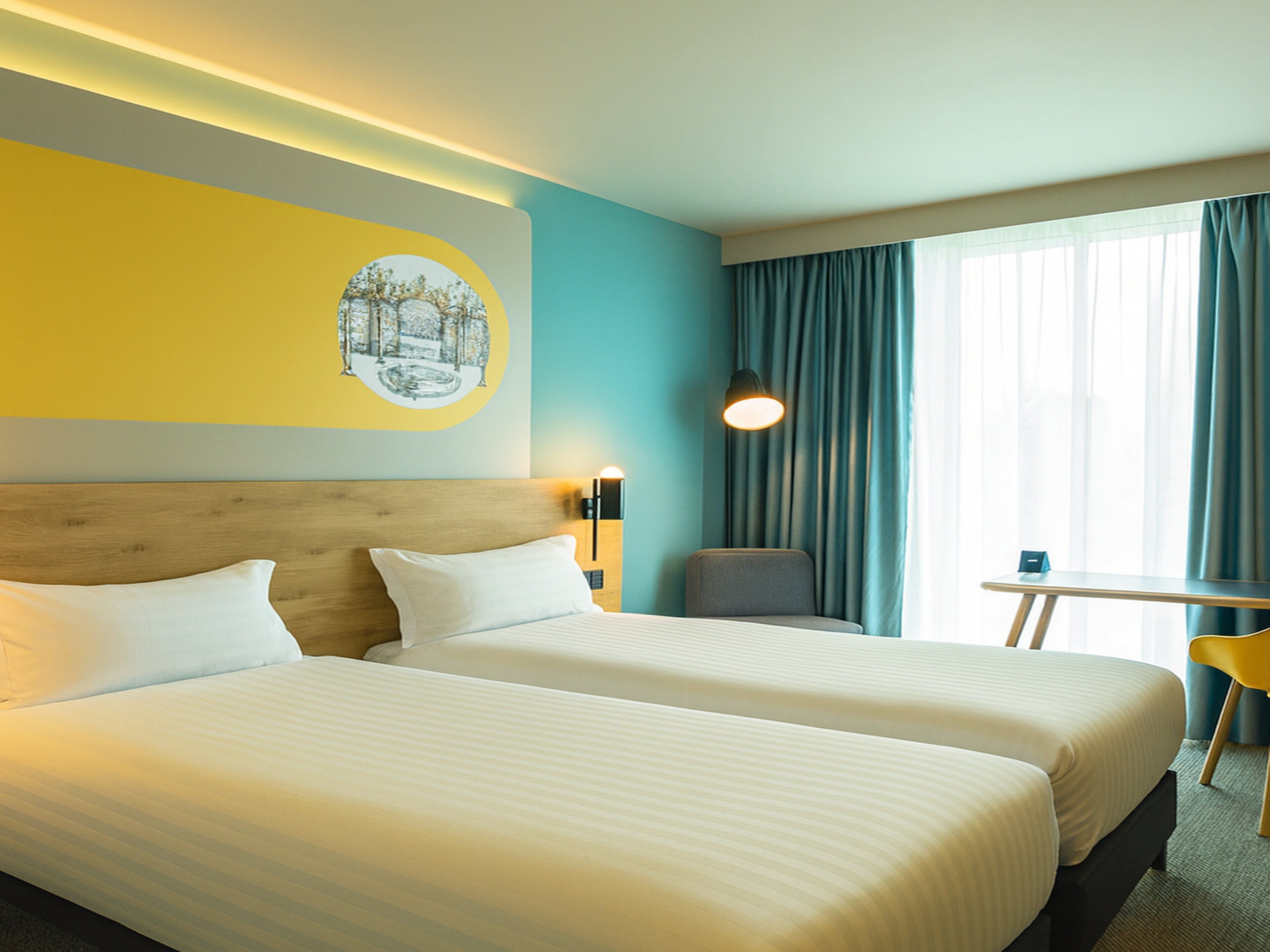 Habitación de hotel con dos camas, una mesita, una silla, una lámpara y una ventana con cortinas.