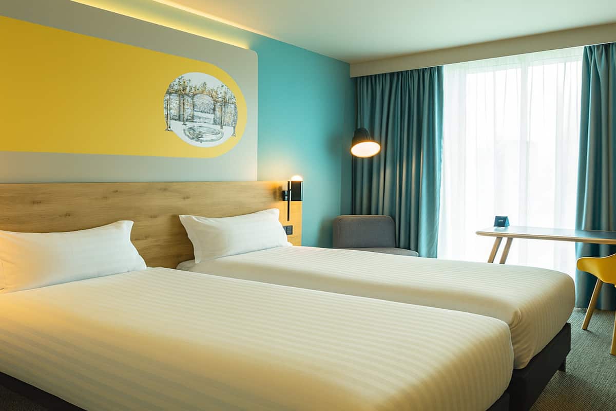Une chambre d’hôtel avec deux lits, une petite table, une chaise, une lampe et une fenêtre avec des rideaux.