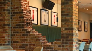 A brick wall with a staircase leading to a green booth area, framed pictures on the wall, and checkered chairs in the foreground. - B&B HOTEL Warrington (Warrington)