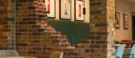 A brick wall with a staircase leading to a green booth area, framed pictures on the wall, and checkered chairs in the foreground.