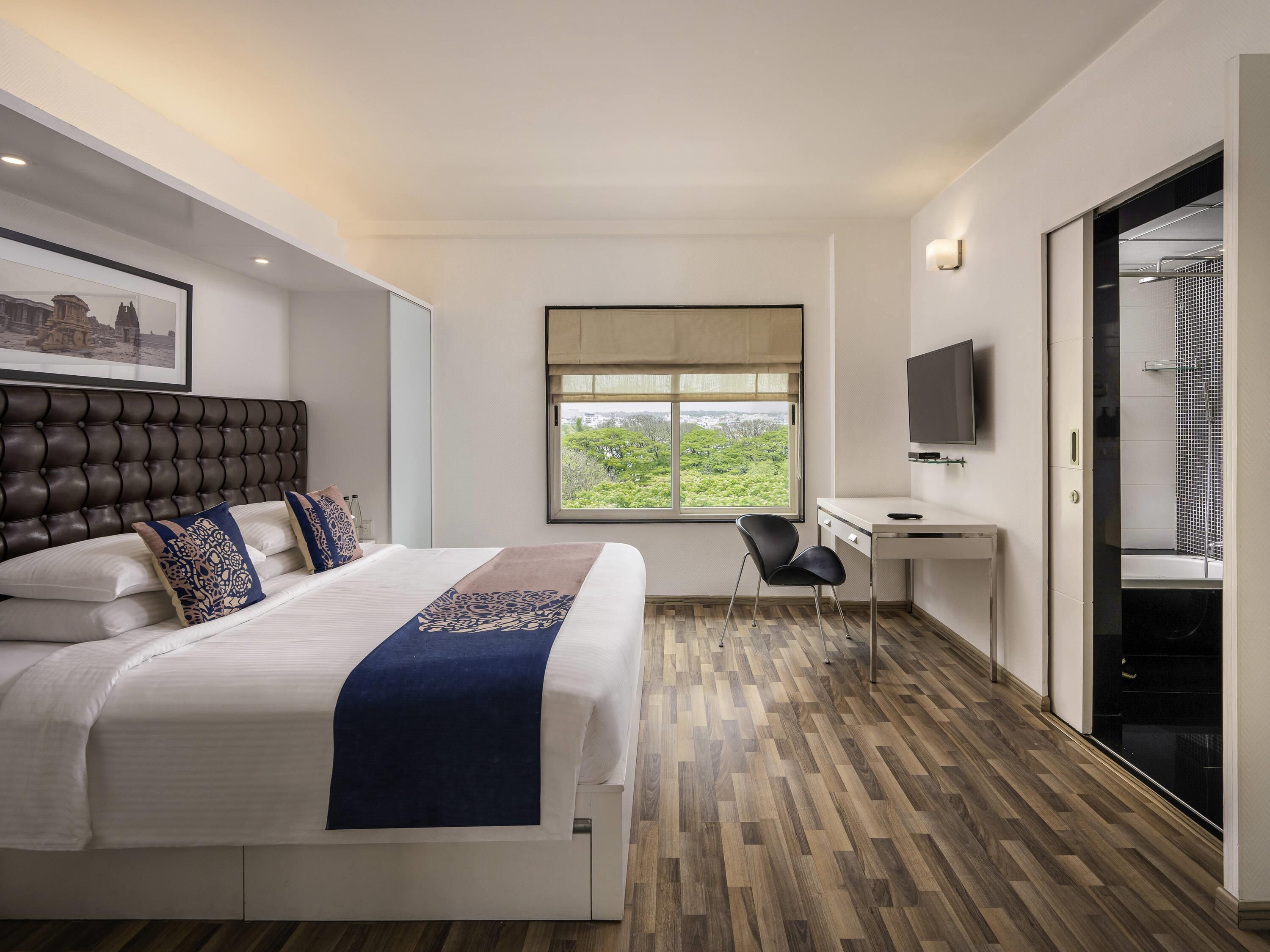 A modern hotel room with a large bed, a desk, a chair, a television, and a window with a view.