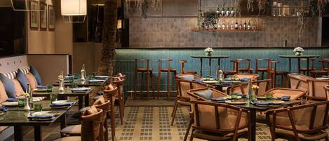 Interior de un restaurante con mobiliario de madera, una zona de bar con estantes llenos de botellas y plantas colgantes.