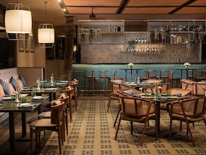 Interior de un restaurante con mobiliario de madera, una zona de bar con estantes llenos de botellas y plantas colgantes.