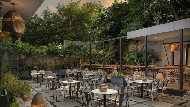 A modern outdoor cafe with round tables and chairs, surrounded by lush greenery and a concrete wall. - Grand Mercure Bangalore (Bengaluru)
