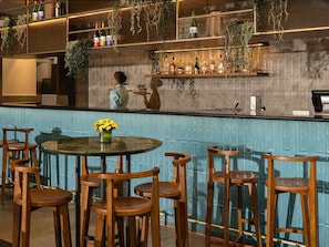 A bar area with wooden stools, a round table with a vase of yellow flowers, and shelves with bottles and plants. - Grand Mercure Bangalore (Bengaluru)