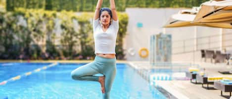 A person performing a yoga pose by a swimming pool with a wooden deck and lounge chairs in the background.