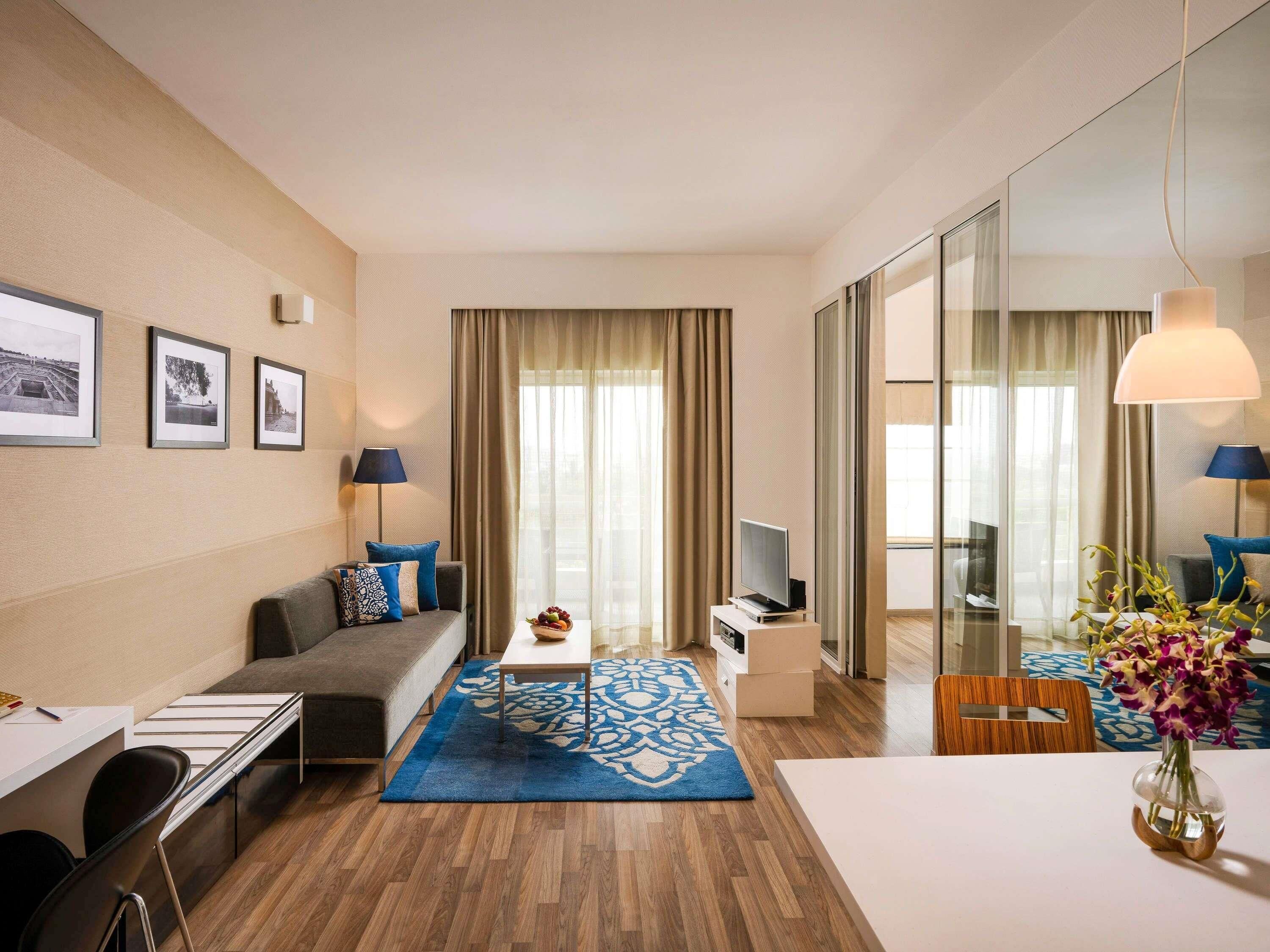 Ein modernes Hotelzimmer mit einer Couch, einem Esstisch, einem Fernseher und einem großen Fenster mit Vorhängen.