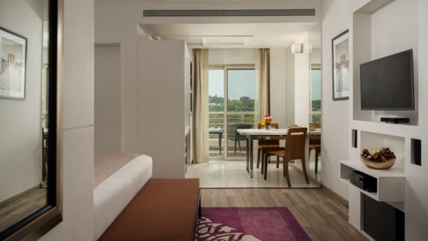 A modern hotel room with a bed, a bench, a dining area with a table and chairs, a TV mounted on the wall, and a balcony with a view.
