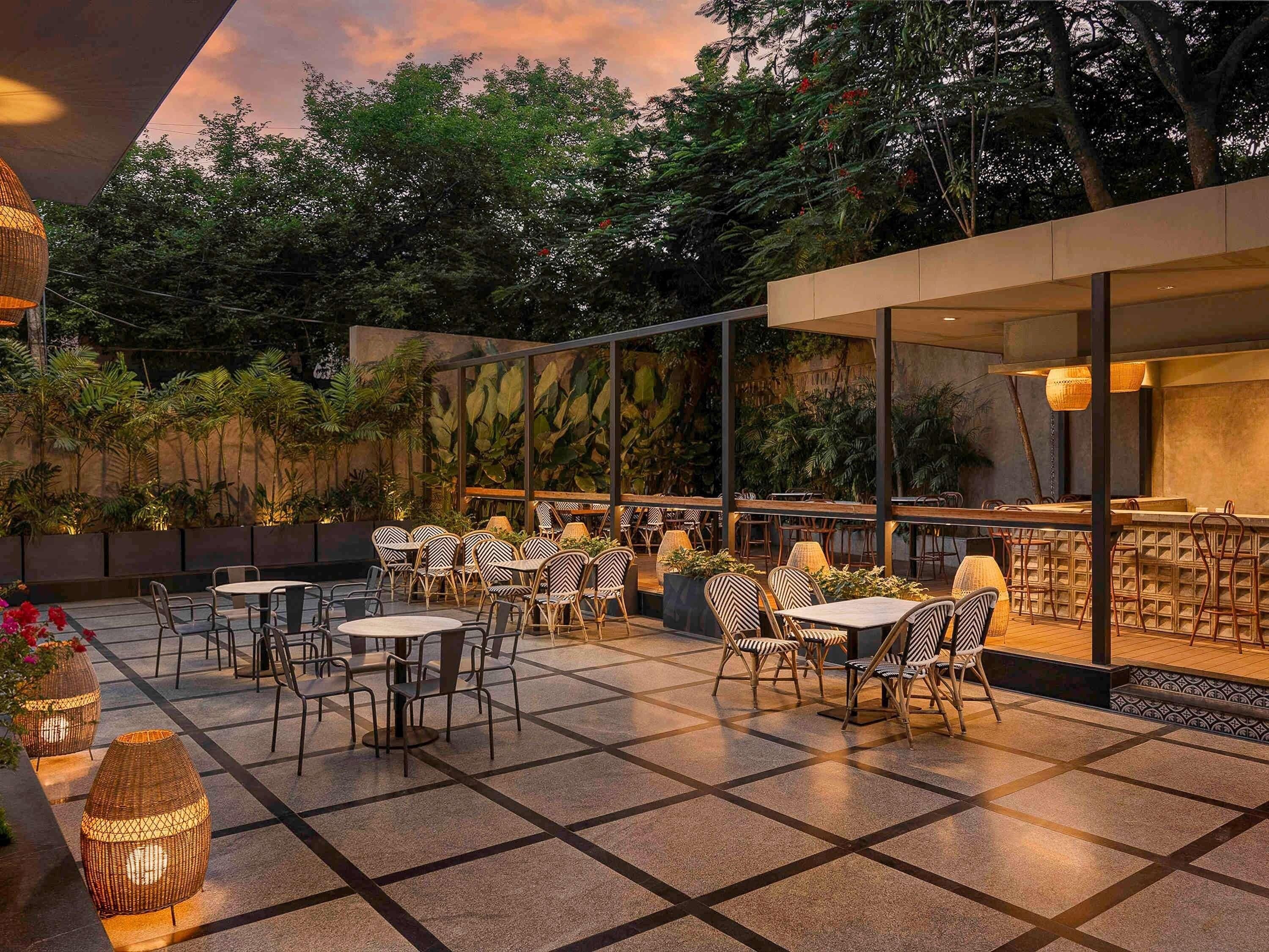 An outdoor patio with tables and chairs, surrounded by greenery and a wall with a mural.