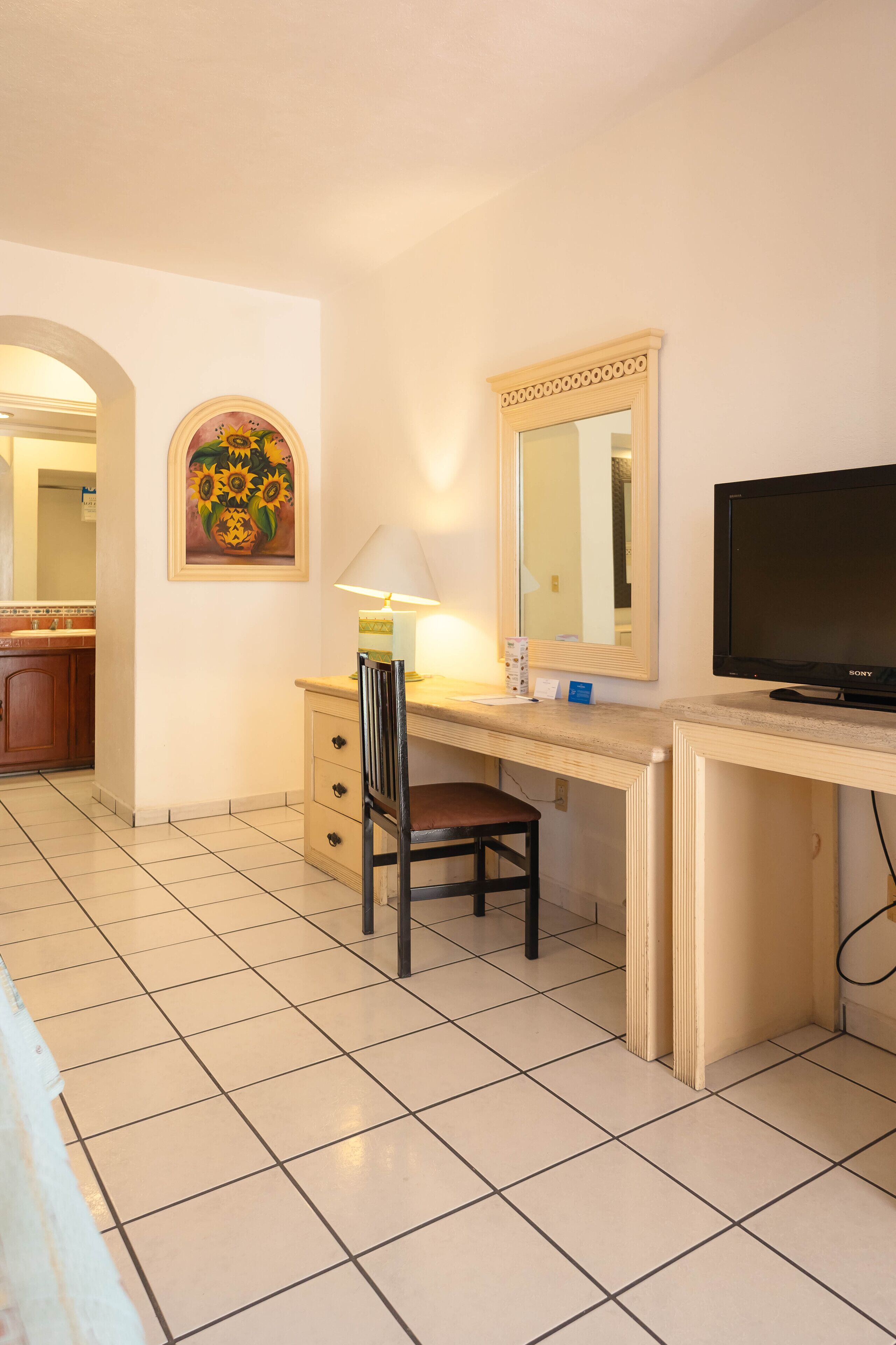 Habitación de hotel con escritorio, silla, televisor, lámpara y un cuadro.